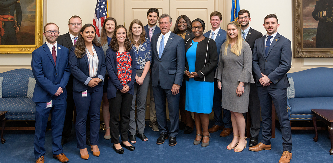 Graduate and undergraduate students stand in professional business attire with John Carney, in the Governor's Office